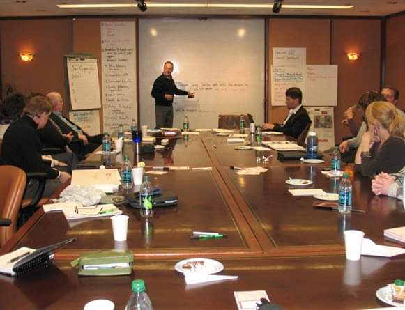 Trey Malicoat facilitating a group learning session around a conference table, with a whiteboard and flip charts.